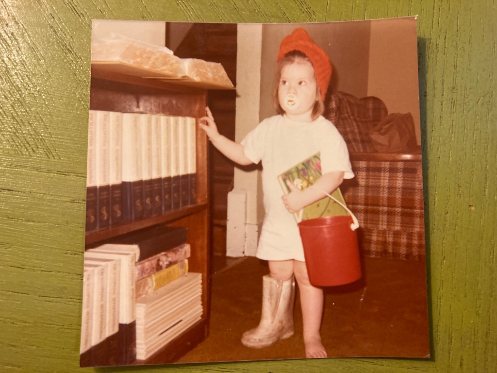 Photo of a toddler in a late 1970s living room, dressed in an oversized white t-shirt, red hat, and one dirty snow boot. She is holding a bucket (like a purse) and a picture book. She looks like she thinks she’s ready to go somewhere.